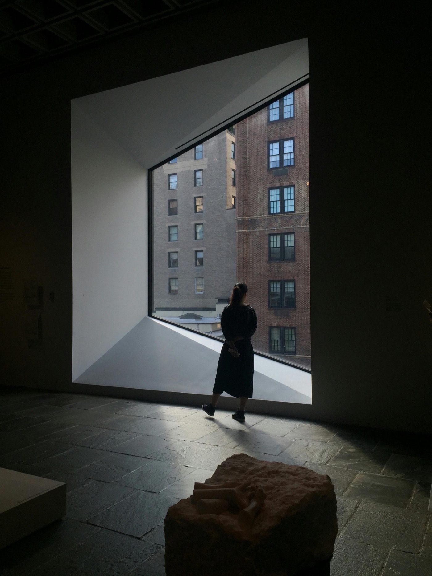A figure standing in a quiet gallery framed by a large rectangular window overlooking a New York City building